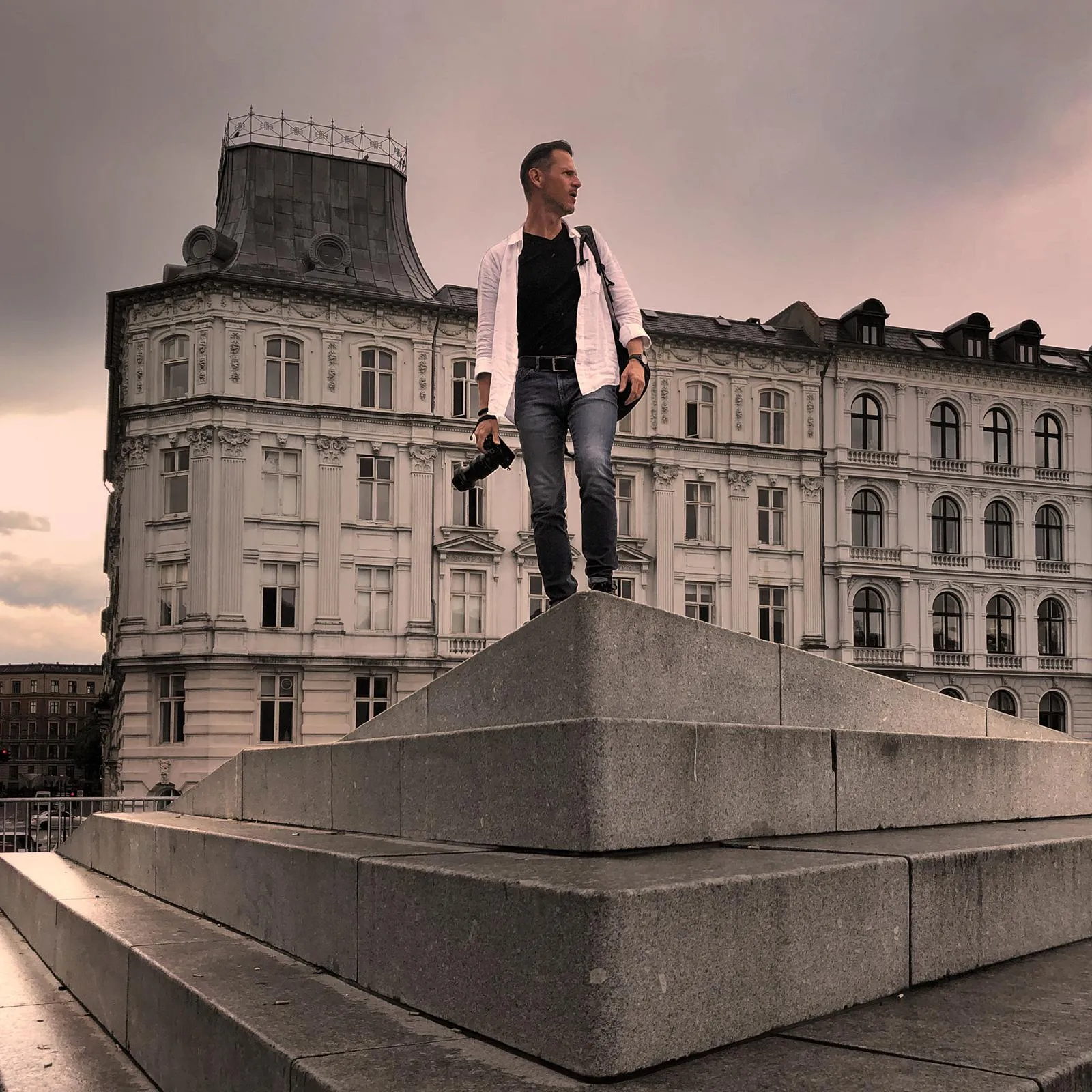 Milan Vodrážka standing atop stone steps holding a camera, with Copenhagen architecture in the background