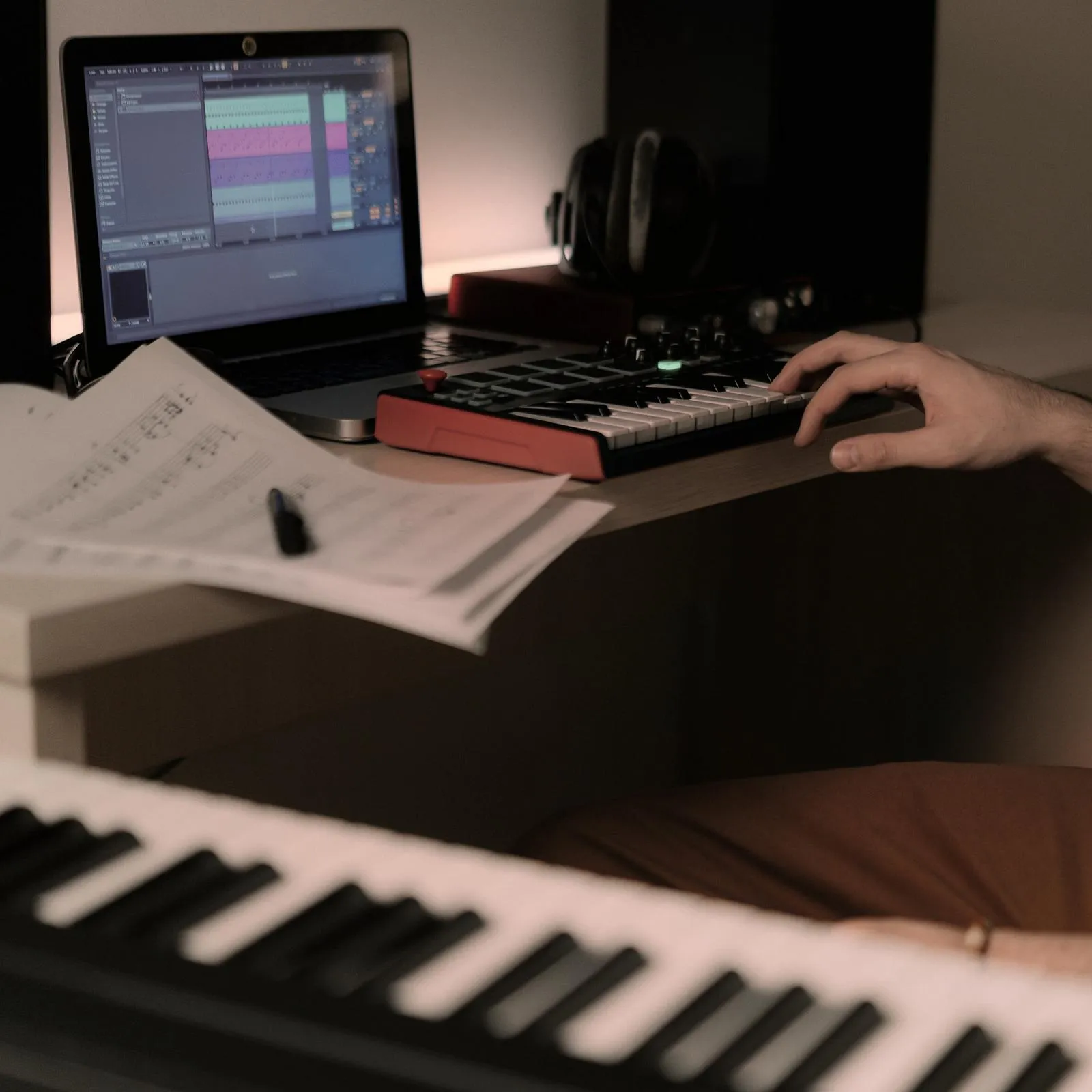 MIDI keyboard beside a laptop running music production software, with handwritten sheet music and a piano in the foreground