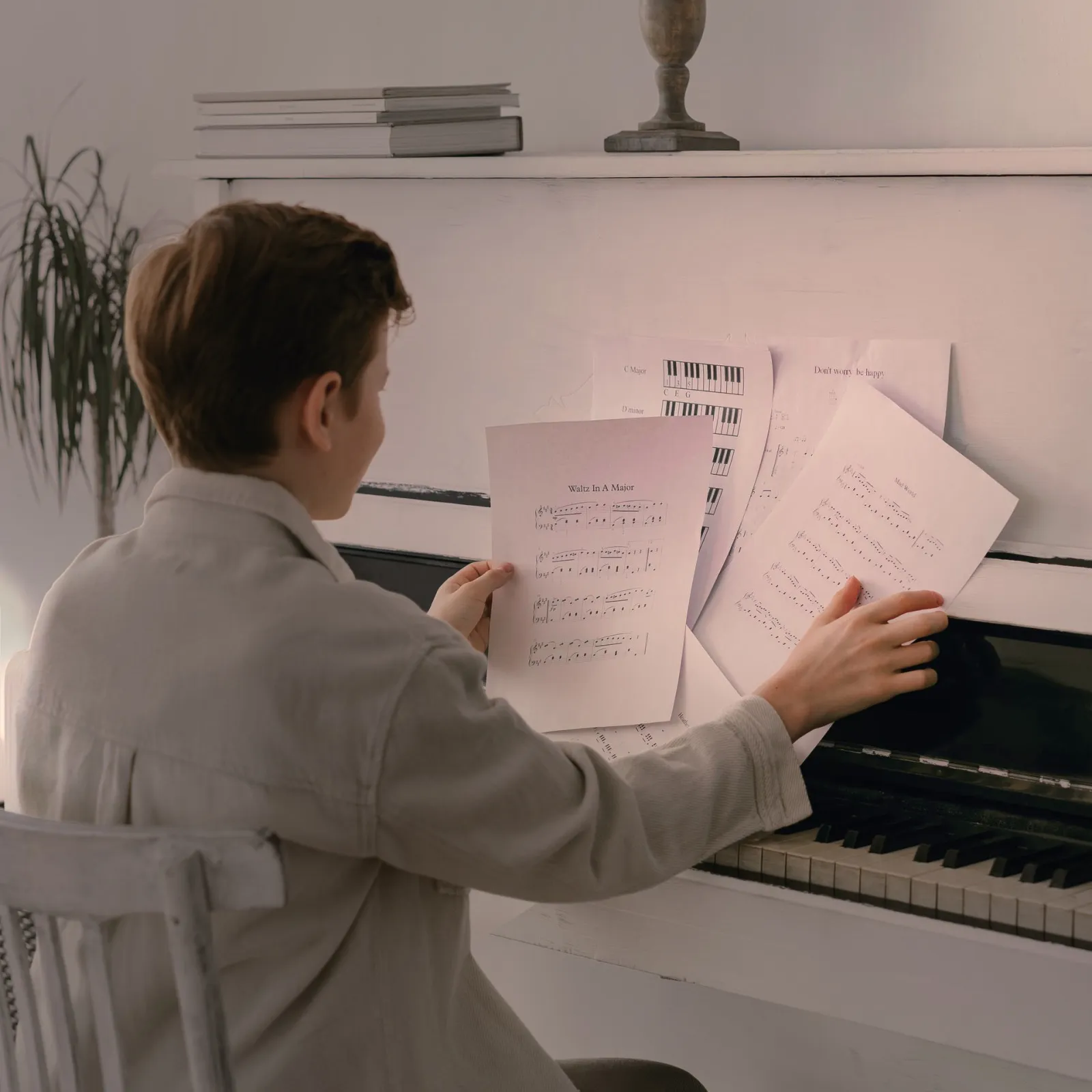 Child seen from behind at a white piano, holding a sheet of music, more pages spread across the piano in front of them